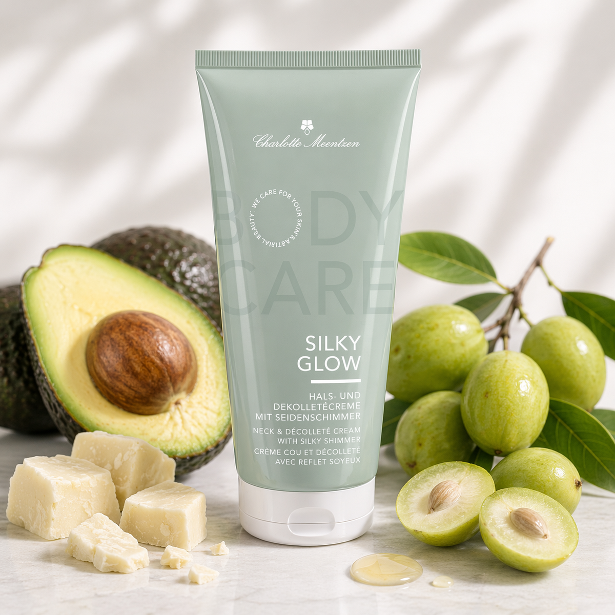 Creme-Tube vor einer aufgeschnittenen Avocado, grünen Früchten und Sheabutter auf hellem Hintergrund.