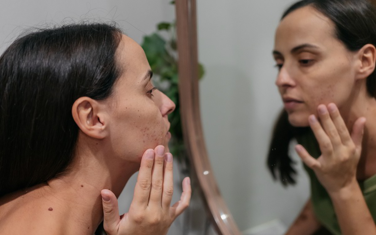 Eine Frau betrachtet sich im Spiegel und berührt nachdenklich ihr Gesicht.